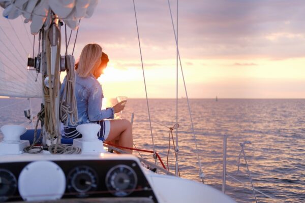 a woman holding a drink and looking down at sunset, sitting on the front of her sailboat with the horizon behind her.